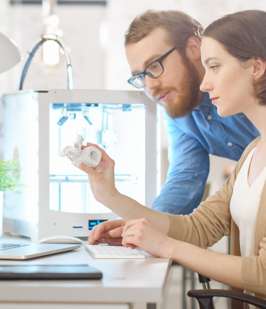 Shot of a Young Bearded Male Manager Helping to a Female Engineer Developer, who is using Laptop Computer and Expecting 3D Model. Bright Modern Office with Programmable 3D Printer.