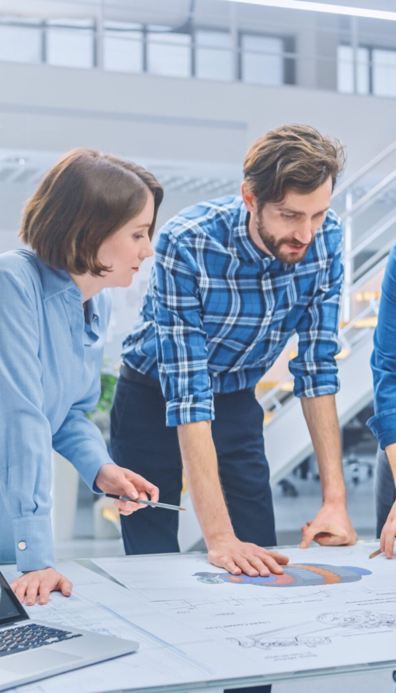 In the Industrial Engineering Facility: Diverse Group of Engineers and Technicians on a Meeting Gather Around Table with Engine Design Technical Drafts, Have Discussion, Analyse Technology